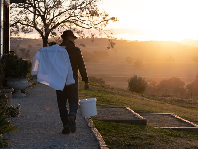 man walking towards sunset with bucket and shirt over shoulder